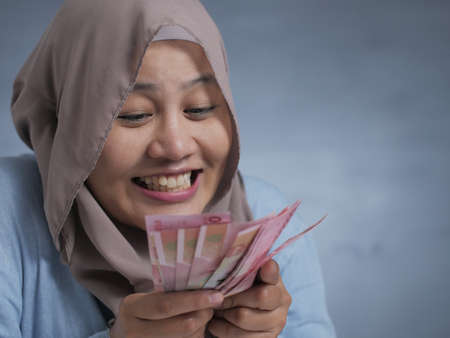 Portrait of Indonesian muslim woman holding rupiah money, smiling laughing winning gesture