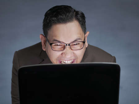 Portrait of young funny Asian man smiling when looking at his laptop, mysterious cunning laugh,...