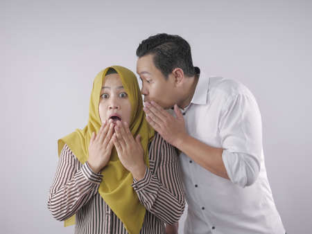 Man covering his mouth and whispering secret to his wife ear, Asian muslim woman looking shocked and...