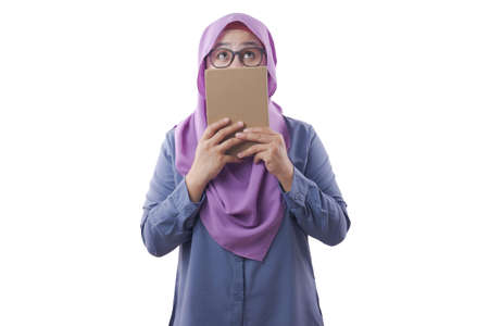 Portrait of young Asian muslim woman wearing hijab smiling and holding book with thinking expression