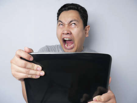 Young Asian man wearing casual shirt looking at laptop, angry gesture. Bad news or failure in...