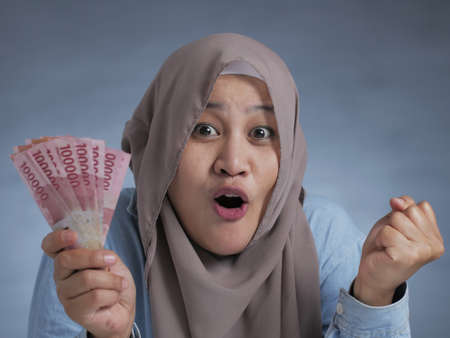Portrait of Indonesian muslim woman holding rupiah money, smiling laughing winning gestureの写真素材