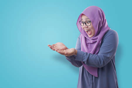 Portrait of Asian muslim woman wearing hijab shows copy space empty palm presenting something, excited smiling expression over blue backgroundの写真素材