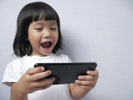 Portrait of little Asian girl looks happy and excited to play with smart phone gadget, bad habit too young with gadget conceptの写真素材