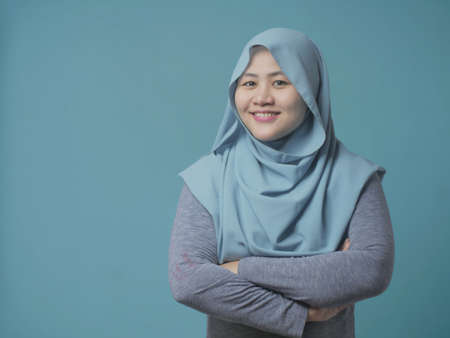 Asian muslim woman wearing blue shirt and purple hijab smiling friendly with arms crossed, confident successful woman standing over blue backgroundの写真素材