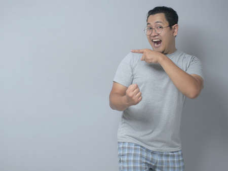 Portrait of funny young Asian man smiling and pointing to presenting something on his side, against grey background with copy spaceの写真素材