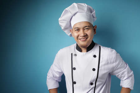 Portrait of happy proud Asian chef smiling at camera, against blue backgroundの写真素材