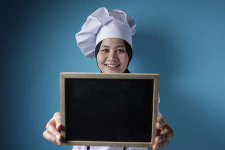 Portrait of Asian female chef looks happy and proud showing empty blackboard, presenting blank writing template, copy space meal menu conceptの写真素材
