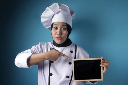 Portrait of Asian female chef looks happy and proud showing empty blackboard, presenting blank writing template, copy space meal menu conceptの写真素材