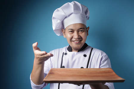 Portrait of Asian male chef looks happy and proud presenting something on his empty wooden plate, copy space meal menu conceptの写真素材