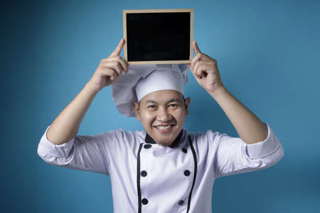 Portrait of Asian male chef looks happy and proud showing empty blackboard, presenting blank writing template, copy space meal menu conceptの写真素材