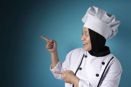 Portrait of Asian female chef looks happy and proud pointing something on her side, cpresenting opy...