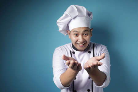 Portrait of happy Asian chef smiling when presenting something on his empty hands, copy space, against blue backgroundの写真素材