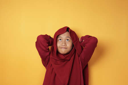 Portrait of young Asian muslim teenage girl wearing red hijab looking up and thinking against yellow backgroundの写真素材