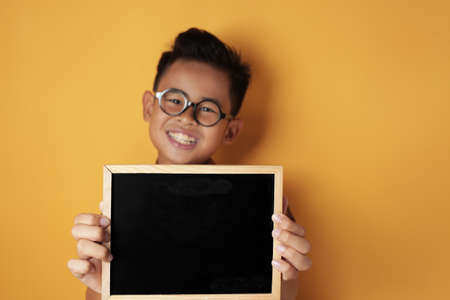 Portrait of smart young cute Asian boy smiling at camera while holding and showing empty blackboard or chalk board against yellow background, copy spaceの写真素材