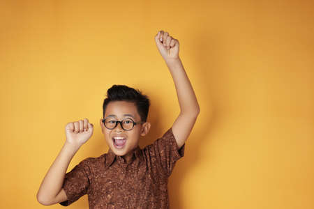 Portrait of cute smart young Asian boy wearing eyeglasses and batik shirt shows winning gesture and smiling at camera, celebrating success victory against yellow backgroundの写真素材