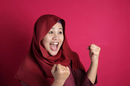 Portrait of success beautiful Asian muslim woman wearing hijab showS winning gesture, excited happy expression celebrating victory, against red backgroundの写真素材