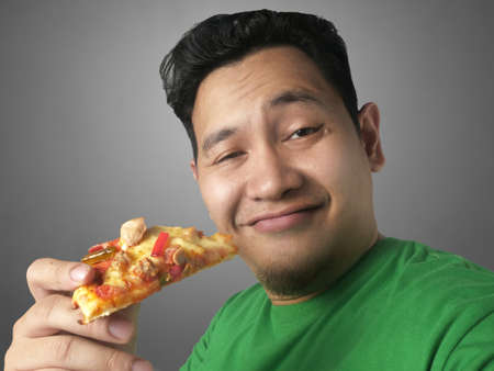Close up portrait of young funny Asian man eating a slice of pizza, unhealthy eating habit, junk...