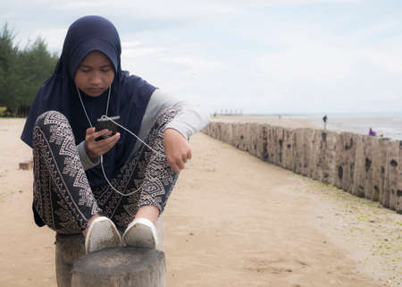 Portrait of Asian teenage muslim girl listeing to music on phone, sitting on the beachの写真素材