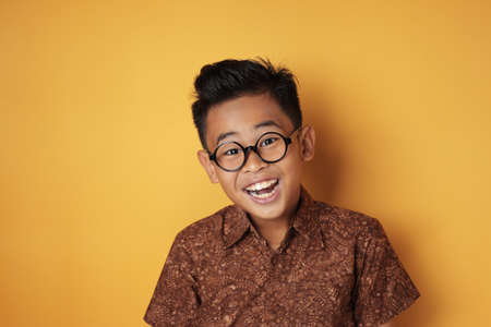 Portrait of young aute Asian boy wearing eyeglasses and batik shirt looking at camera and laugh, against yellow backgroundの写真素材