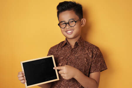 Portrait of smart young cute Asian boy smiling at camera while holding and showing empty blackboard or chalk board against yellow background, copy spaceの写真素材