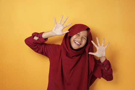 Portrait of happy Asian muslim teenage girl smiling and waving at camera, wearing red hijab against yellow backgroundの写真素材