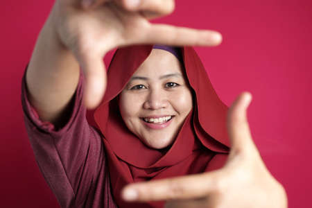 Portrait of happy Asian muslim woman smiling at camera while framing her face with hands, against red backgroundの写真素材