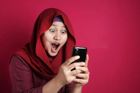 Portrait of young Asian muslim woman get good news on her phone, happy surprised expression, against red backgroundの写真素材