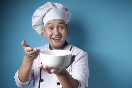 Portrait of Asian male chef looks happy and proud presenting something on her empty white bowl, copy space meal menu conceptの写真素材