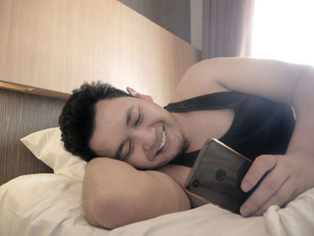 Asian man smiling happily to see good news on message in his smart phone when waking up on bed in the morningの写真素材