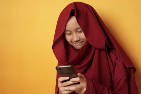 Portrait of young Asian muslim teenage girl with smart phone, smiling happily when reading online chat messaging on her phone against yellow backgroundの写真素材