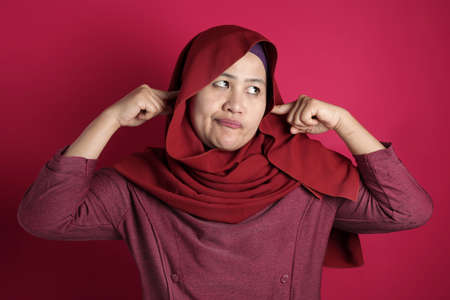 Portrait of Asian muslim lady wearing hijab close hear ears with hands as if do not want to hear something bad, against red backgroundの写真素材