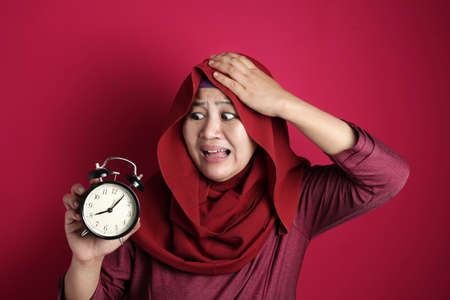 Portrait of muslim woman looked worried afraid while holding and looking at alarm clock, time late concept, against red backgroundの写真素材