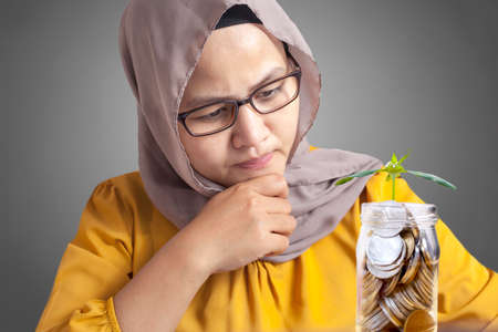 Muslim businesswoman looking at coins in glass jar with plant grows in it, concept of looking for...