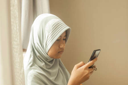 Portrait of young Asian muslim teenage girl with smart phone, smiling happily when reading online chat messaging on her phoneの写真素材
