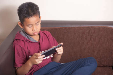 Young Asian boy playing online games on tablet smart phone gadget, sitting on couchの写真素材