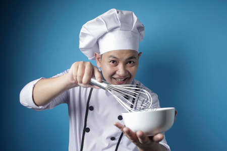 Portrait of Asian male chef smiling happy while holding bowl and whisk, chef mixing ingredient with whisk, against blue backgroundの写真素材