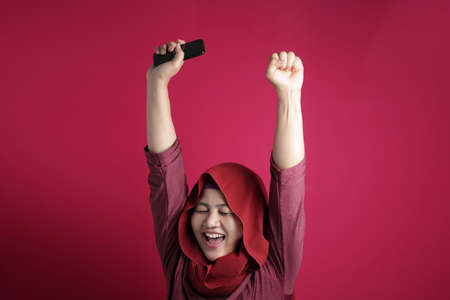 Portrait of young Asian muslim woman get good news on her phone, happy surprised expression, against red backgroundの写真素材