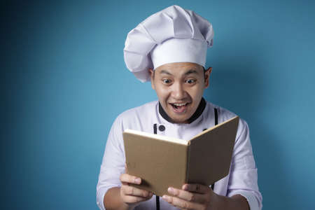 Portrait of Asian male chef reading book of recipes, finding secret recipe, against blue background