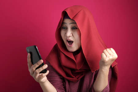 Portrait of young Asian muslim woman get good news on her phone, happy surprised expression, against red backgroundの写真素材