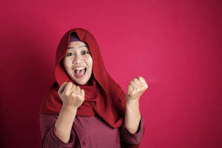 Portrait of success beautiful muslim businesswoman wearing hijab screaming shouting and shows winning victory gesture over red backgroundの写真素材
