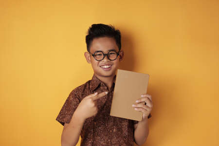 Portrait of Asian student boy holding book and smiling at camera, smart schoolboy, against yellow backgroundの写真素材