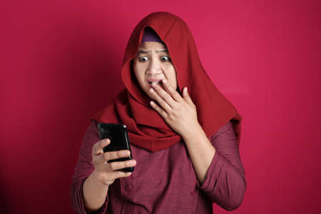 Young Asian muslim woman wearing hijab shows shocked expression, looking at her phone. Close up body portait against red backgroundの写真素材