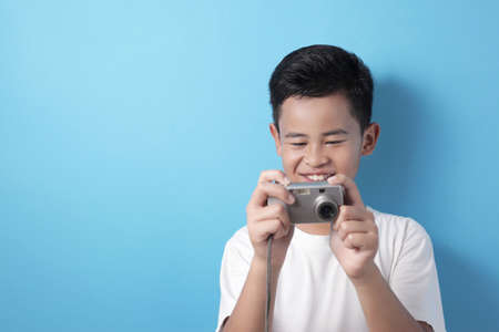 Happy Asian boy doing photography leisure activity, kid as photographer taking picture with his digital camera, against blue backgroundの写真素材