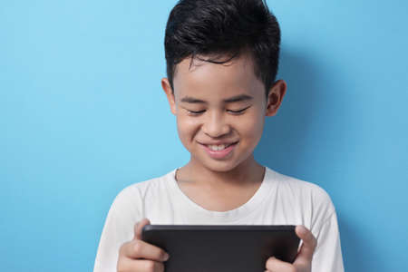 Portrait of Asian boy plays online game on his tablet, smiling happy face, kid with gadget concept