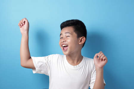 Portrait of happy success smiling Asian boy shows winning gesture, celebrating victory, against blue backgroundの写真素材