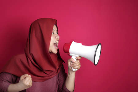 Young Asian muslim businesswoman wearing hijab screaming with megaphone, angry expression. Side view close up body portrait, against red backgroundの写真素材