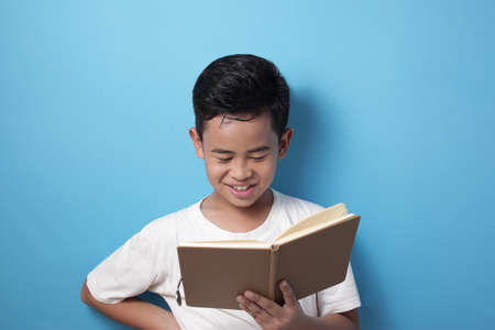 Portrait of happy smart Asian student boy smiling when reading a book, against blue background,...