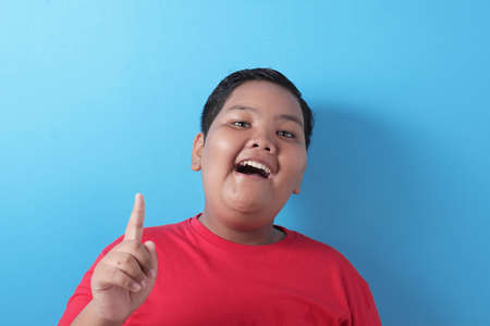 Portrait of happy cheerful fat Asian boy pointing finger up, having idea gesture with big smile, against blue backgroundの写真素材