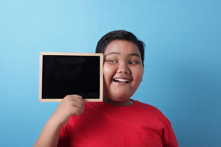 Portrait of healthy fat smart Asian boy smiling at camera while holding and showing empty blackboard or chalk board against blue background, copy spaceの写真素材